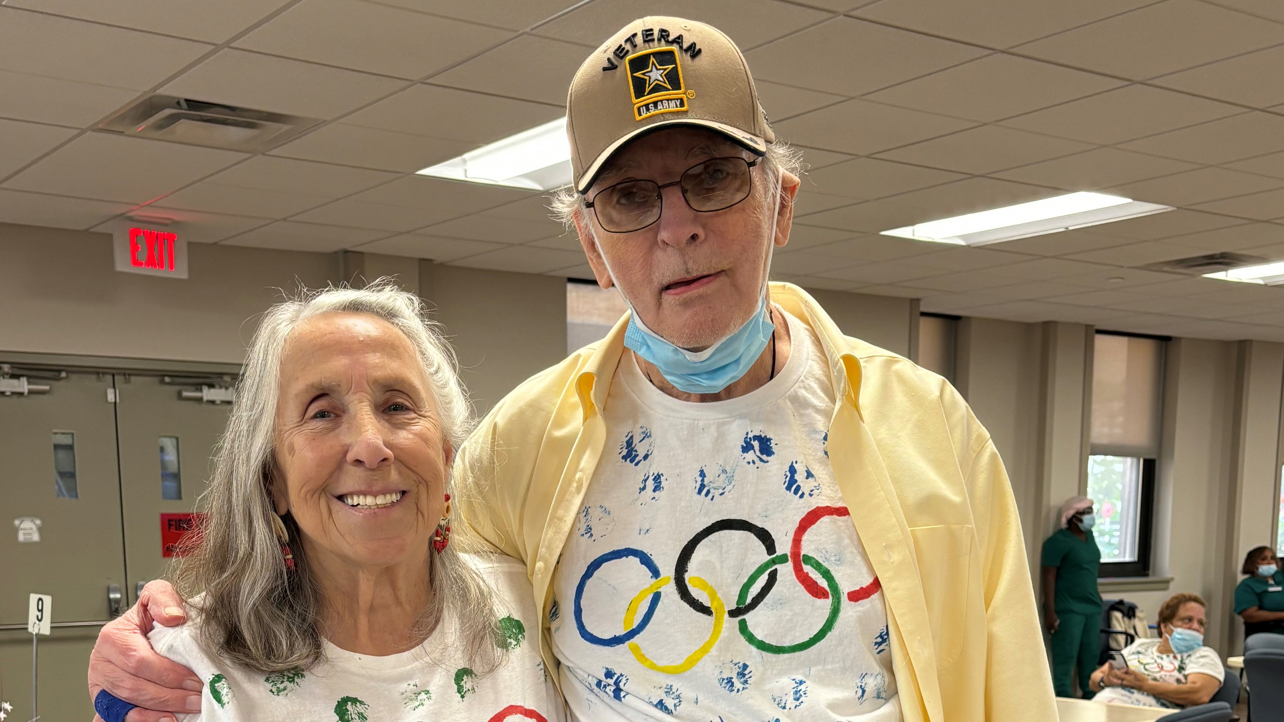 Mr. and Mrs. Goodman stand side by side, each wearing a white T-shirt with the Olympic rings painted on them in blue, yellow, black, green, and red.
