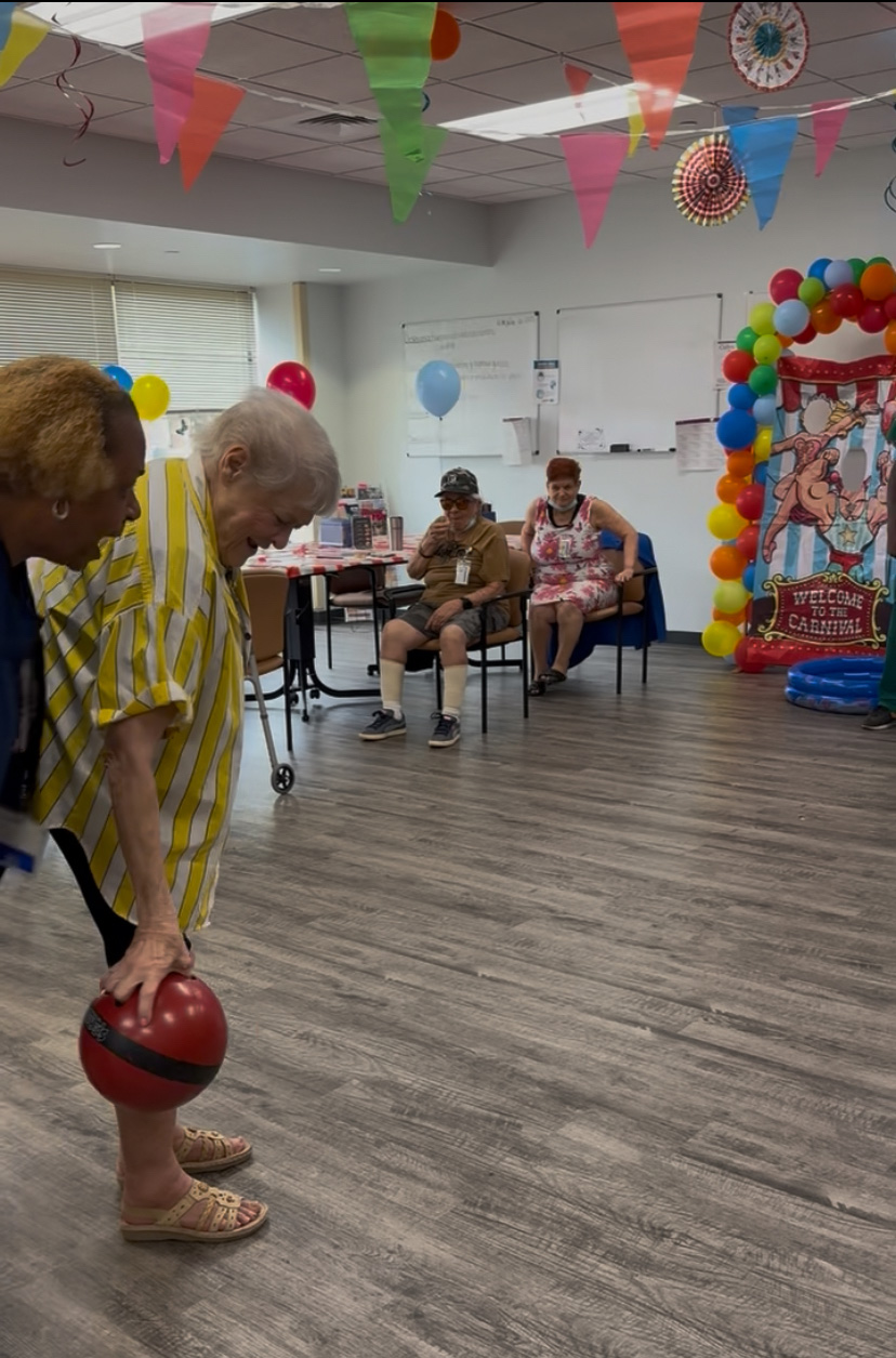 A festive indoor room decorated for a carnival-themed event with colorful triangular flags hanging from the ceiling. A person in a yellow and white striped outfit holds a red and black bowling ball in the foreground. In the background, three people sit near an inflatable carnival game with balloons and a 'Welcome to the Carnival' sign. The room has wood-patterned vinyl flooring and scattered balloons.