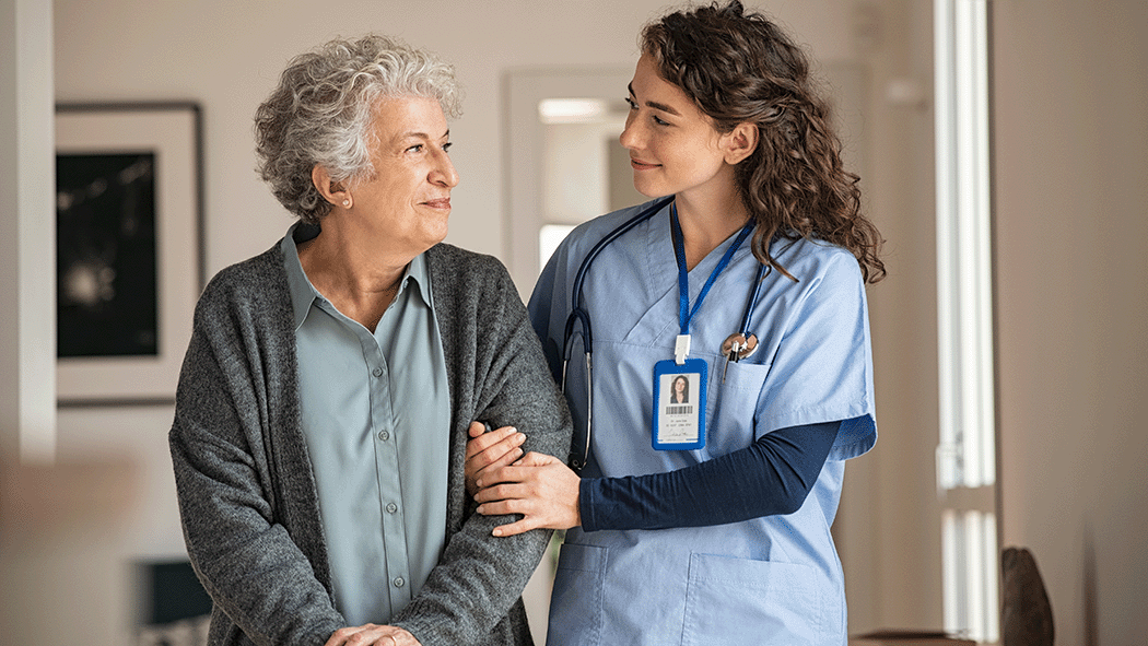 A healthcare worker in light blue scrubs gently supports an older adult by holding their arm as they walk together indoors. The worker has a stethoscope around their neck and a badge visible, offering reassuring assistance.